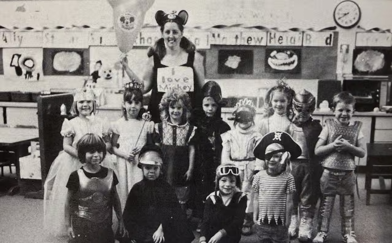 Archival black-and-white photograph of a teacher and children in costumes in a classroom.