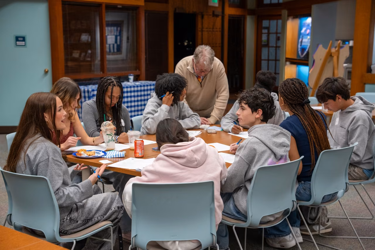 Alumni gather around a library table with a teacher during their reunion dinner.
